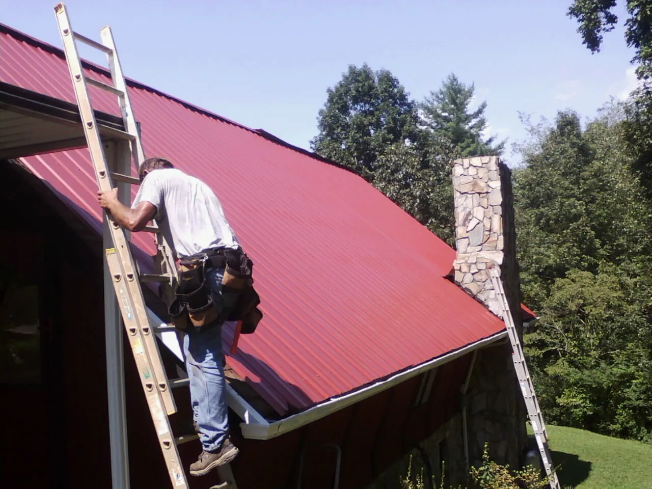 Roofer on a ladder beside a red metal roof and stone chimney — Soffit & Fascia Repair by Roofing Brothers in Excelsior